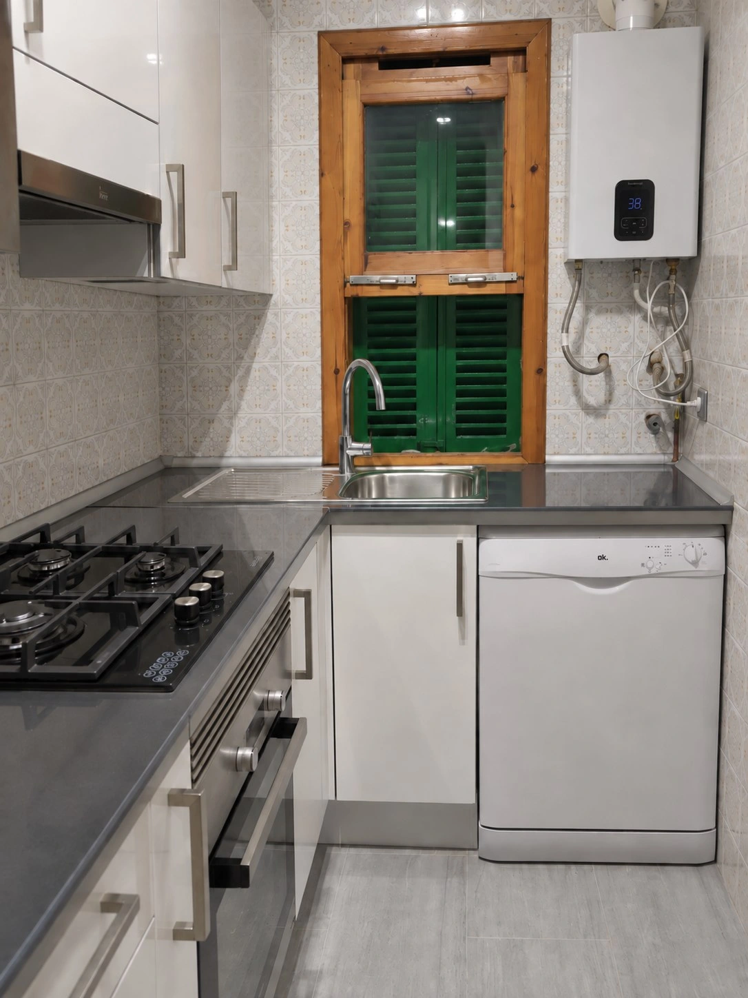 Cocina básica en L terminada: placa de gas integrada en la encimera laminada gris, fregadero bajo la ventana, lavavajillas de libre instalación y las baldosas originales de la pared