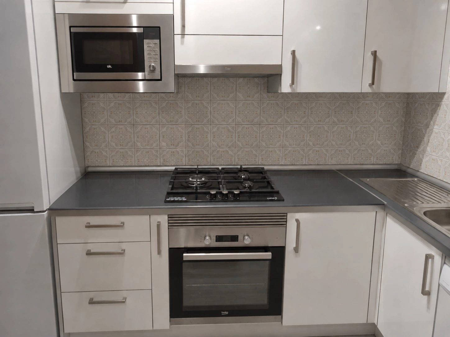 Cocina básica en L en un piso de Mallorca: frentes blancos brillo de catálogo, encimera laminada, placa de gas integrada, microondas encastrado y lavavajillas, con el alicatado original de la pared conservado