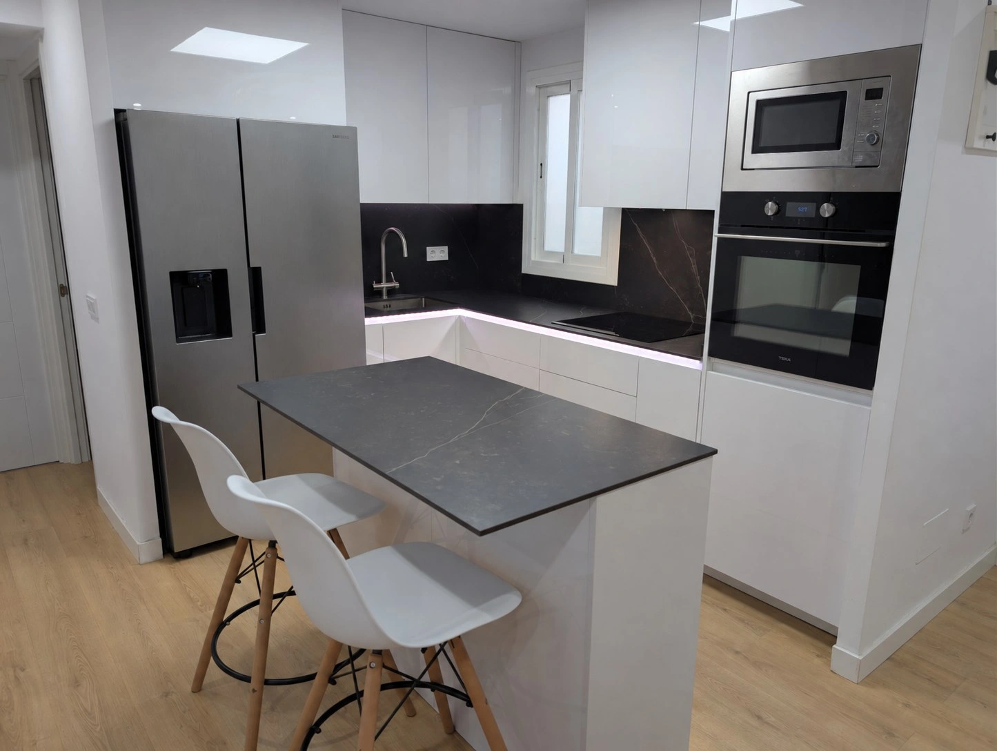Cocina reformada en un piso de Mallorca: isla central con encimera de Dekton y dos taburetes, frigorífico americano en inox, muebles blancos hasta el techo y columna con horno y microondas