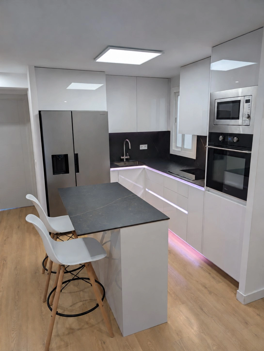 Cocina reformada en un piso de Mallorca con la isla central y sus taburetes, el frigorífico americano en inox y la columna de electrodomésticos al fondo