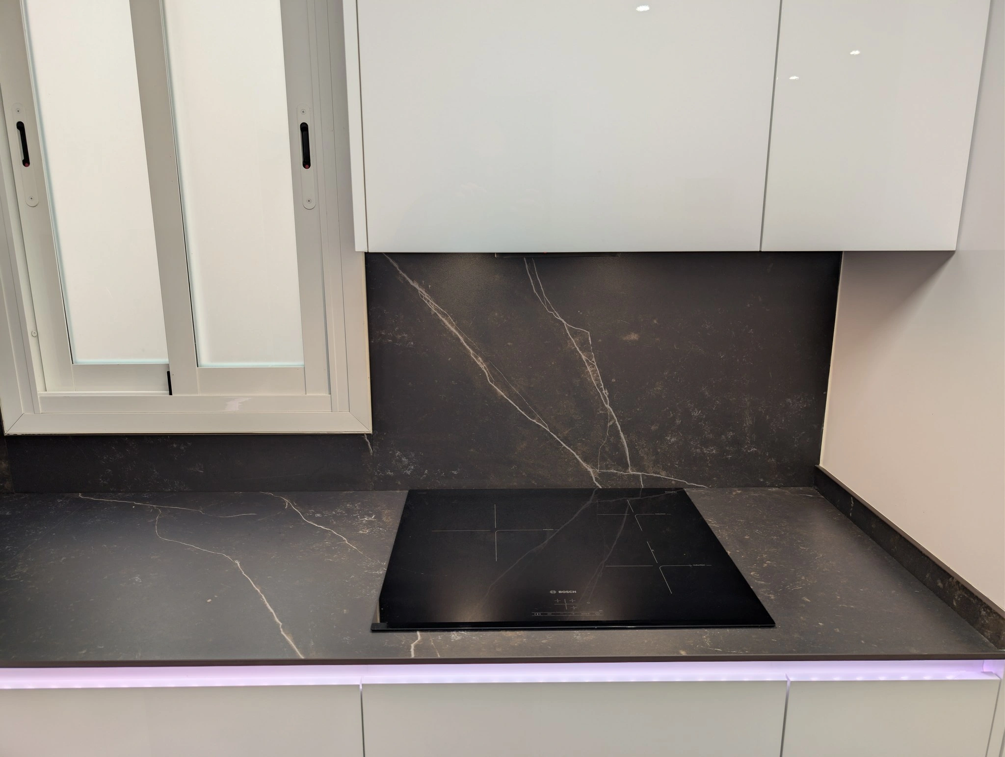 Zona de cocción de la cocina reformada: placa de inducción sobre encimera de Dekton negra, frontal a juego y tira LED encendida bajo los muebles altos